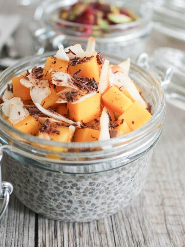 Side view of a clear jar filled with chia seed pudding and toped with diced mango, coconut flakes, and shaved chocolate.