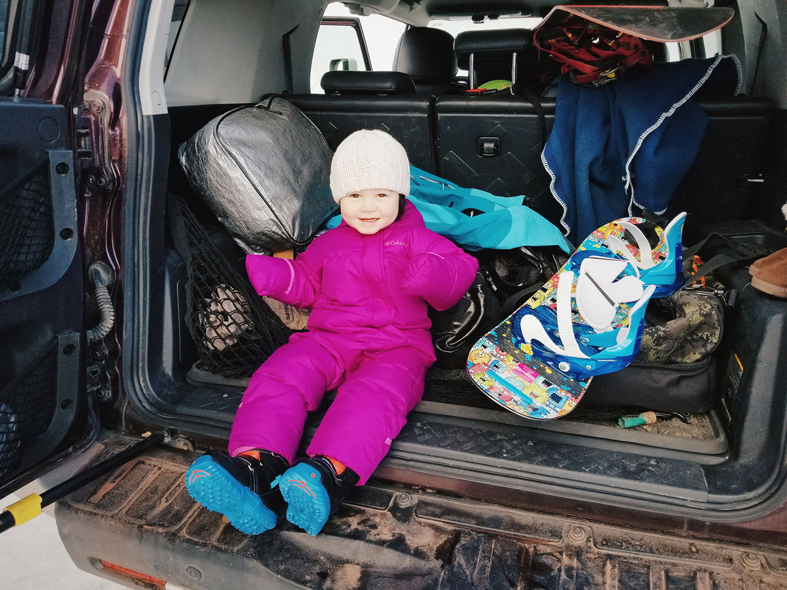 A 2-year-old toddler sitting in the back of a car with her snowboard gear