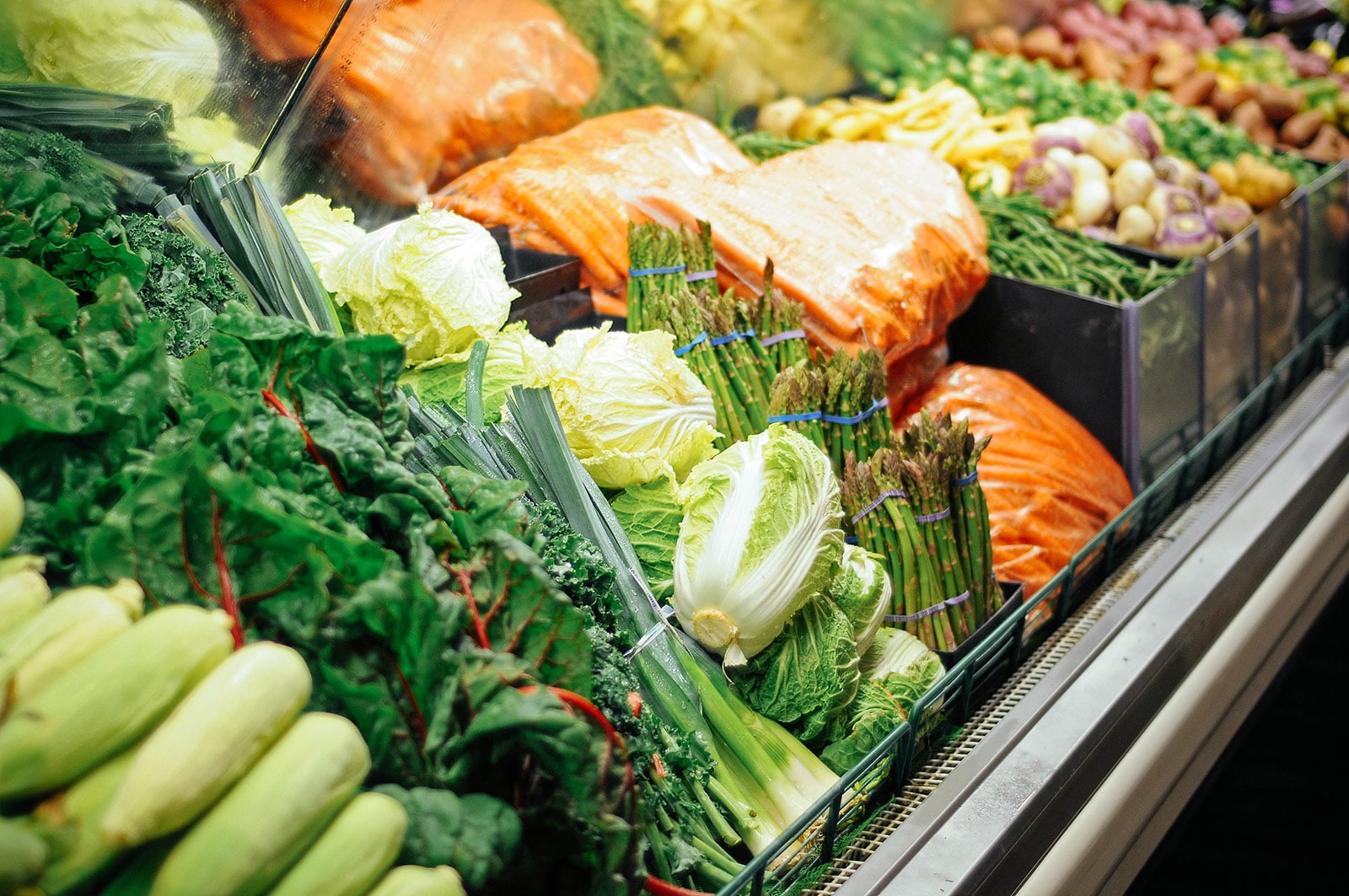 Supermarket produce aisle showing sweet corn, chard, leeks, napa cabbage, asparagus, bags of carrots, and more vegetables in the background