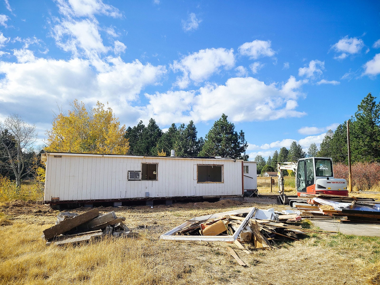 Older mobile home with piles of debris in the yard and a mini excavator parked nearby