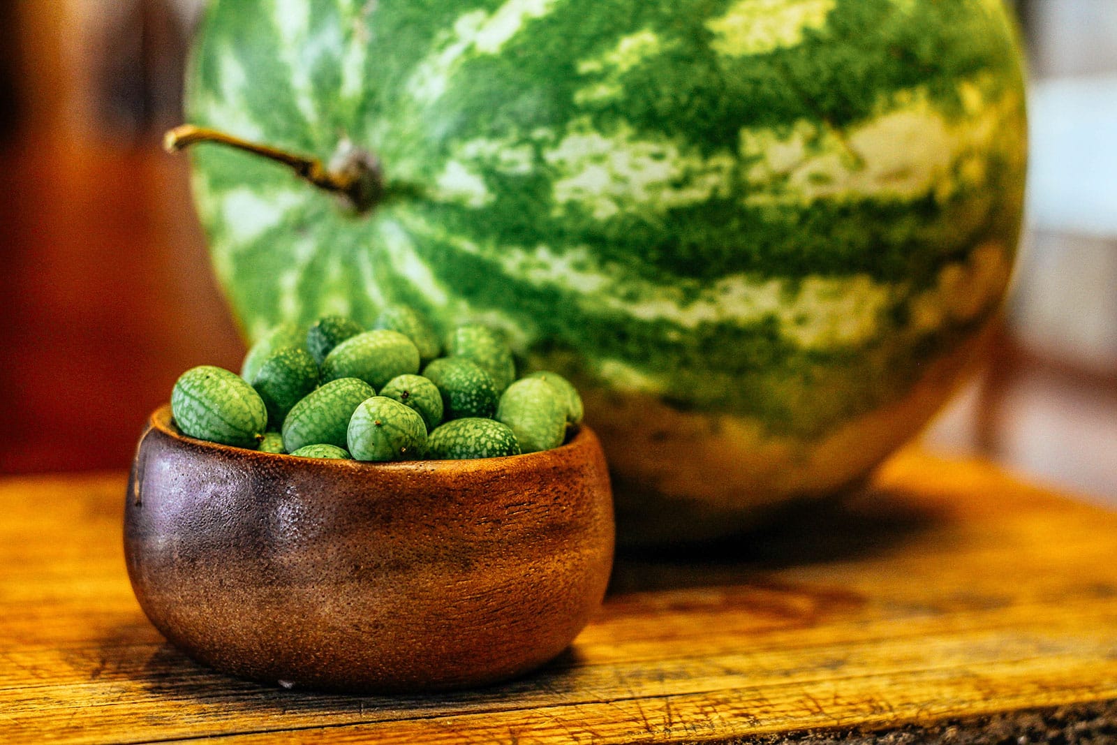 A small wooden bowl filled with cucamelons placed in front of a large watermelon on a wooden surface