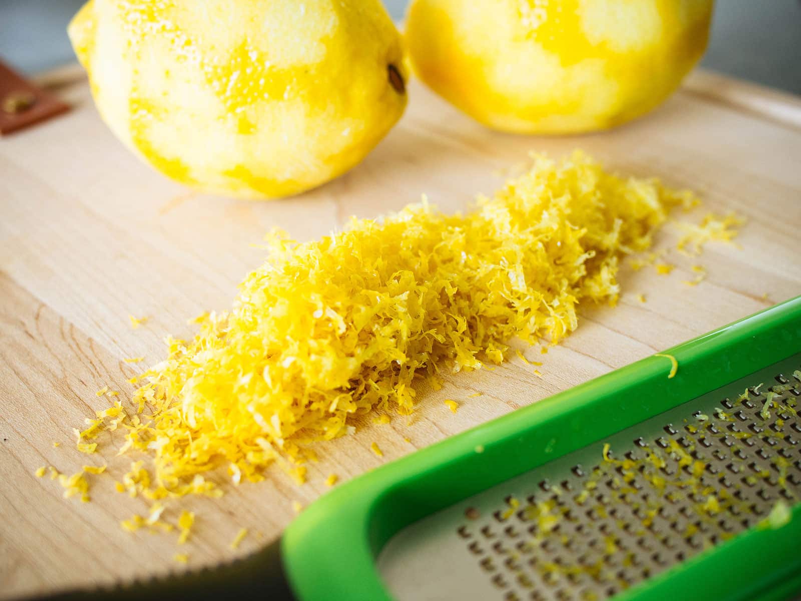 Wooden cutting board with a mound of lemon zest in the center, with peeled lemons in the background and a green Microplane zester in the foreground