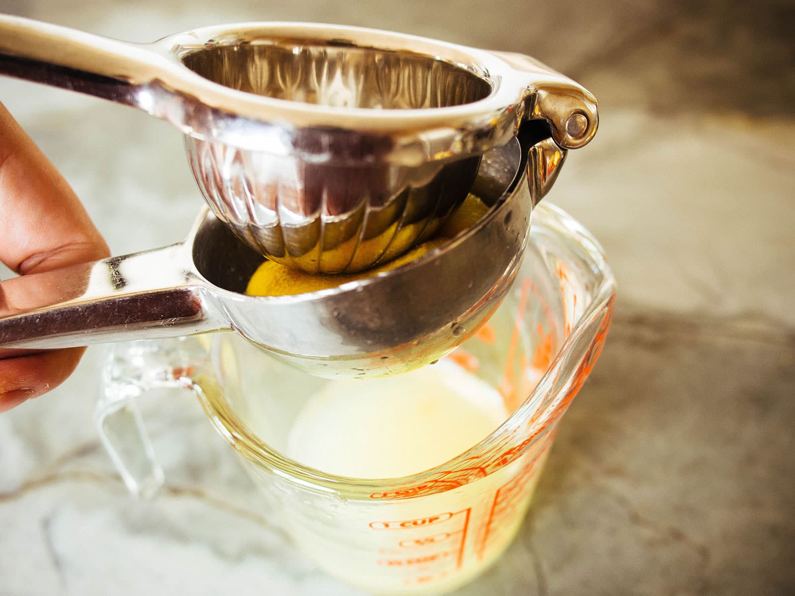 Closeup of a hand squeezing a lemon with a citrus squeezer, and into a glass measuring cup filled with lemon juice