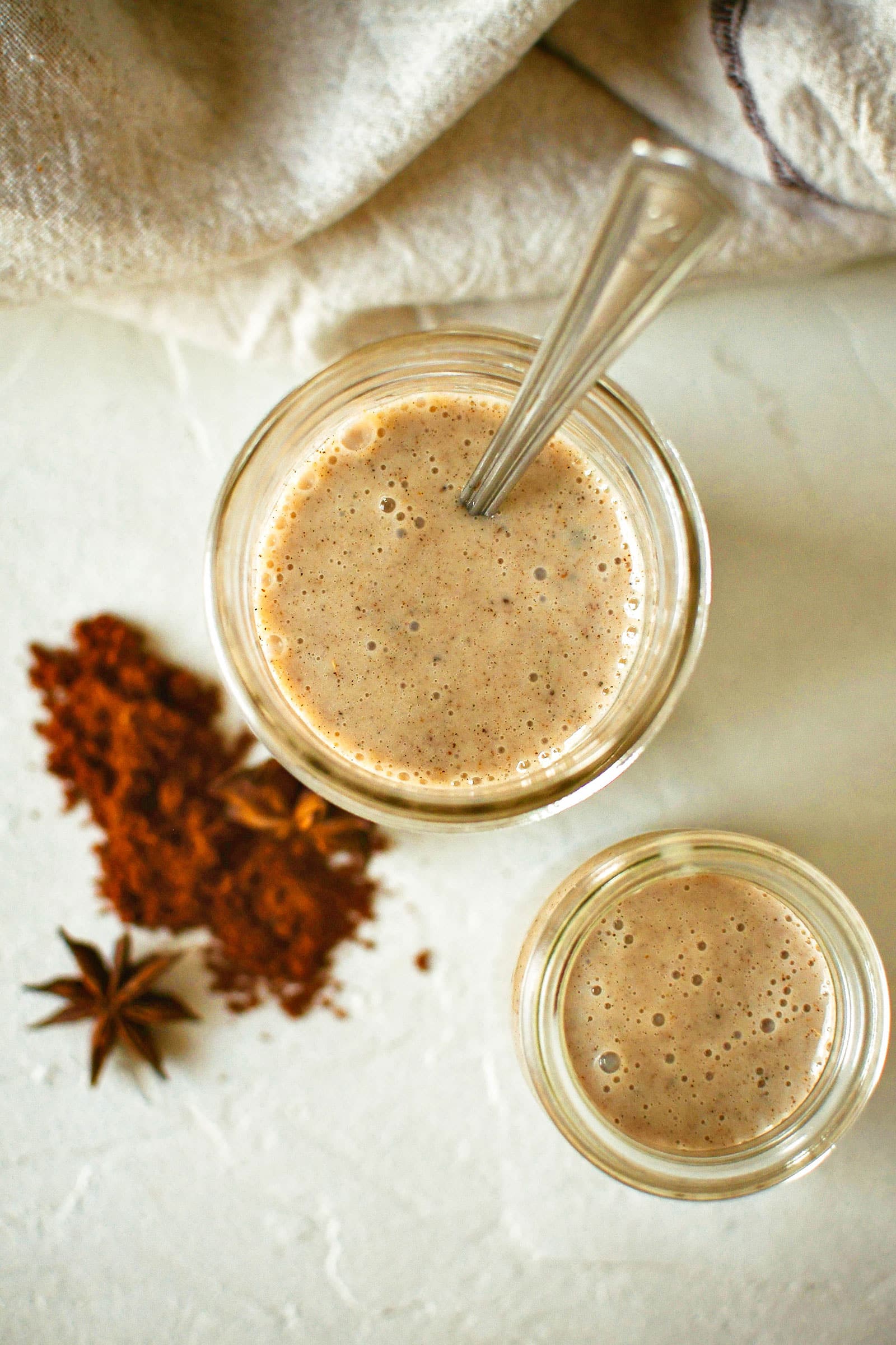 Overhead shot of two jars of homemade chai concentrate shot on a textured white surface with a linen napkin off to the side