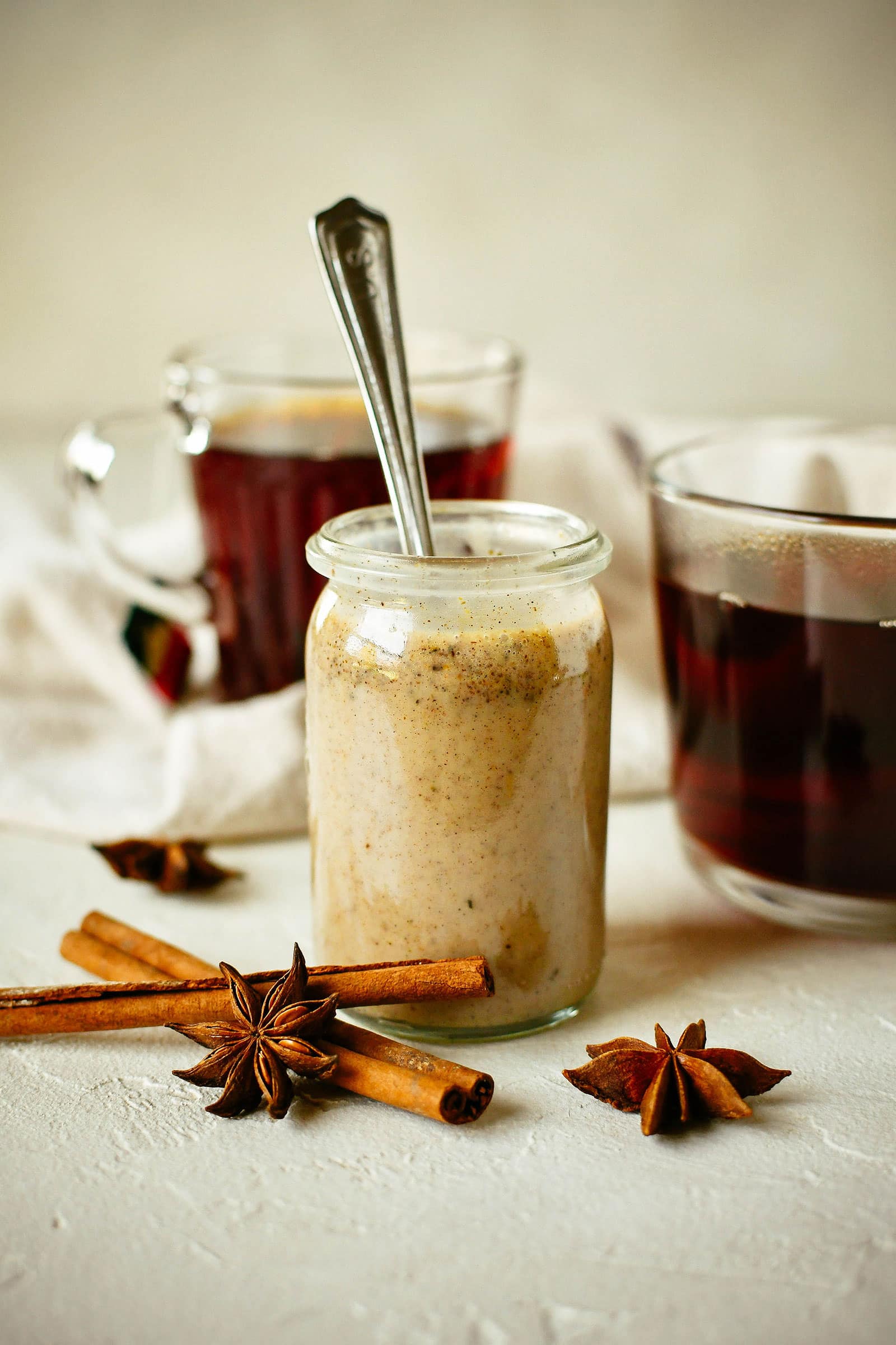 A jar of homemade chai concentrate with a spoon in it, cinnamon sticks and star anise in the foreground, and and mugs of hot tea in the background