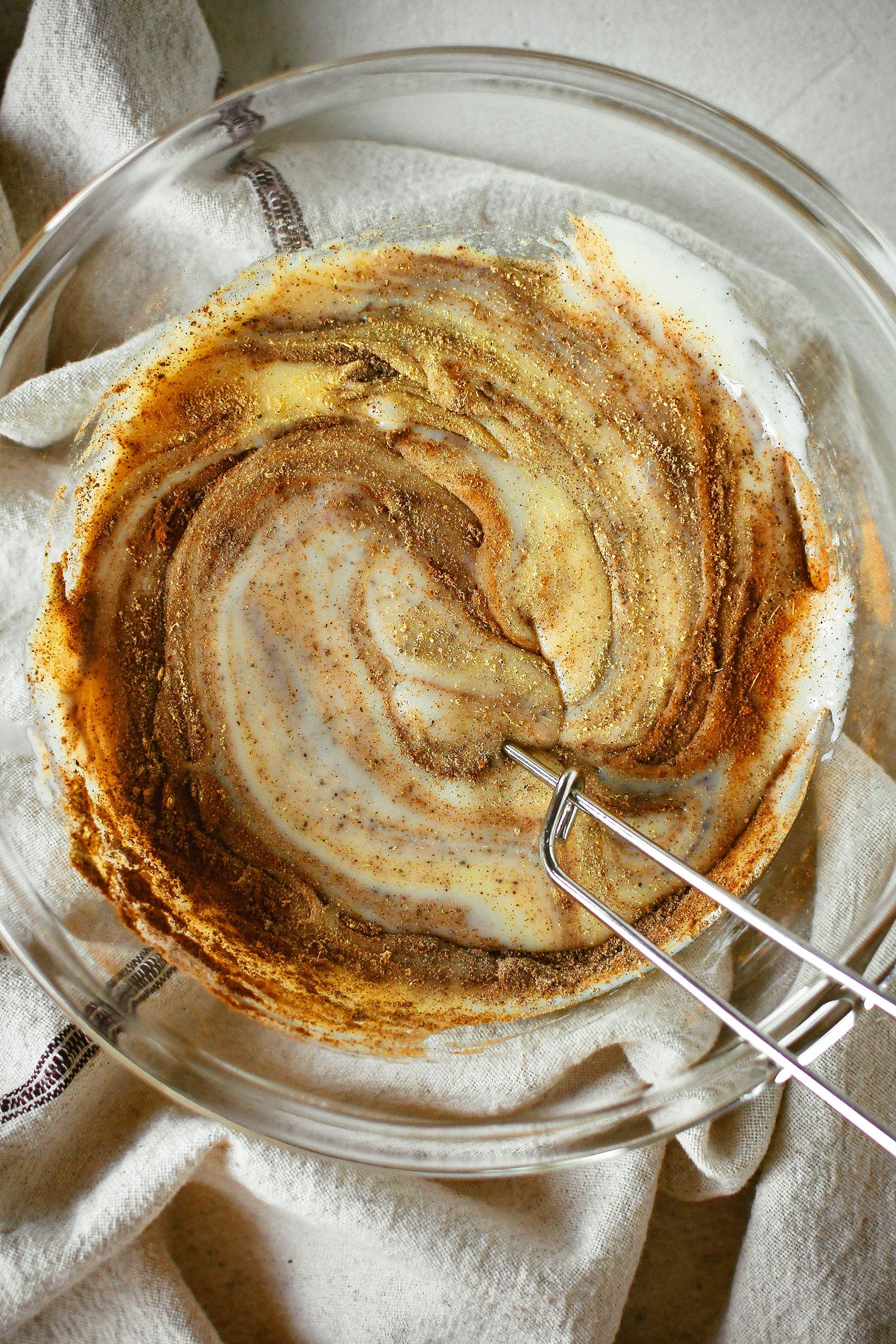 Overhead view of chai spices being whisked into a glass bowl of condensed milk, shot on a linen napkin