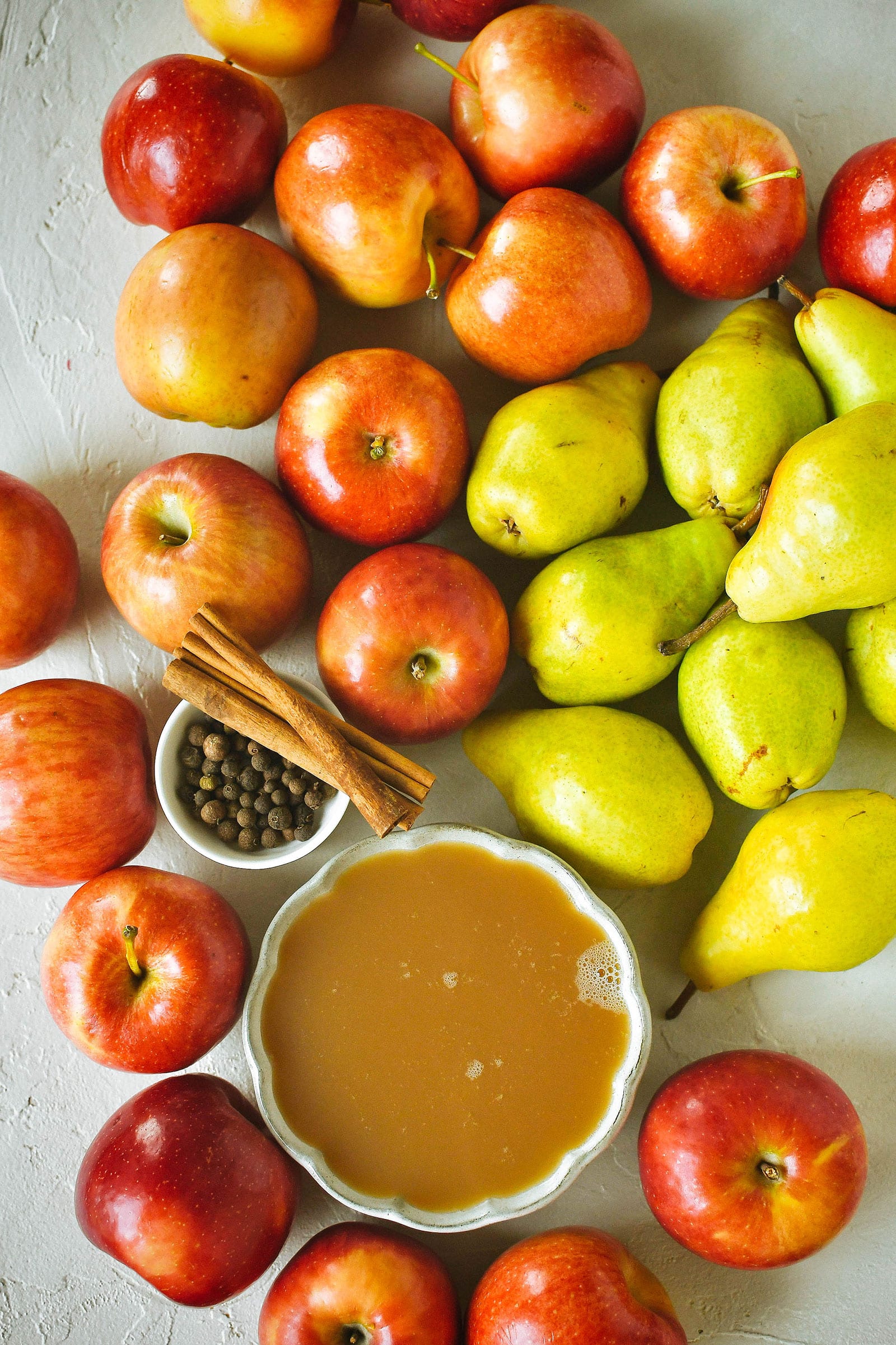 Overhead shot of red apples and green pears, a bowl of apple cider, allspice berries, and cinnamon sticks on a white table