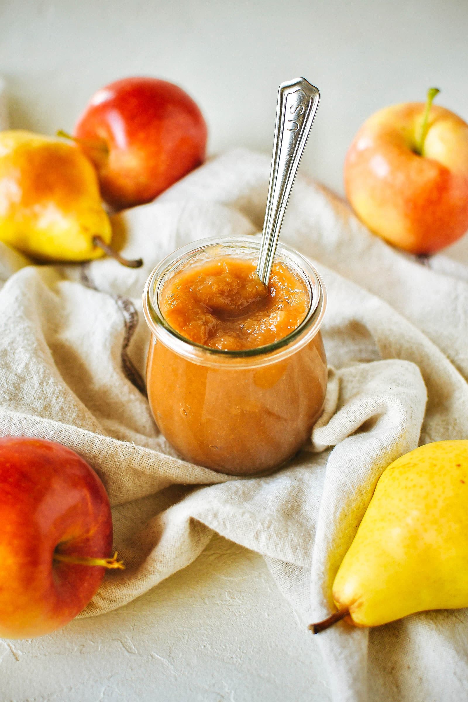 Stylized scene of a half-pint jar of apple-pear butter, with a spoon in the jar and apples and pears arranged around the jar on a linen napkin