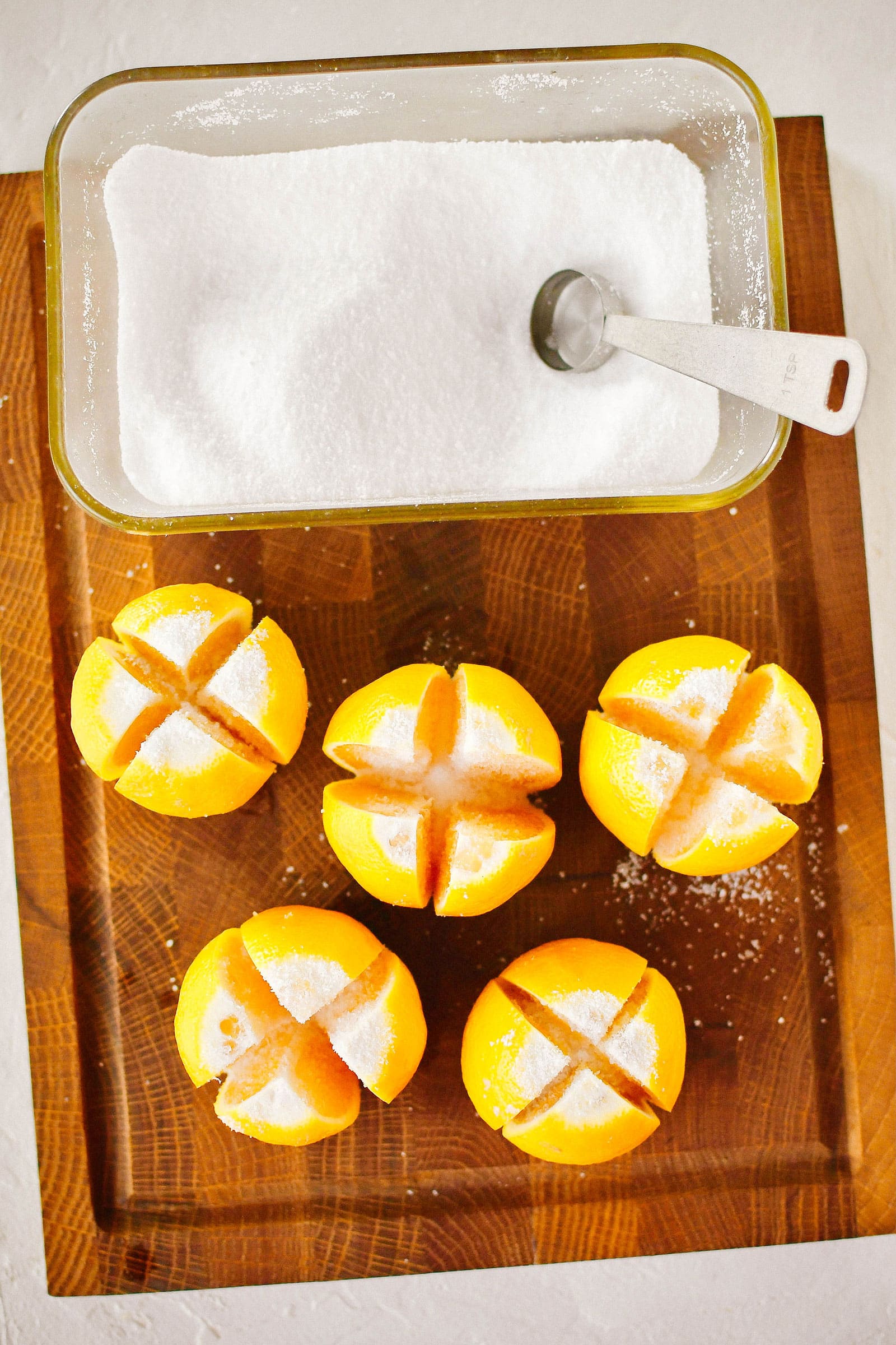 Quartered lemons and a glass container of salt on a wooden cutting board, with each lemon rubbed with salt on the cut sides