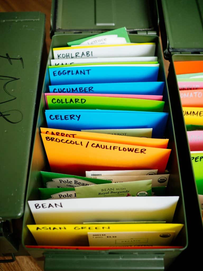 An open metal ammo can revealing packets of seed packets inside, categorized by plant name